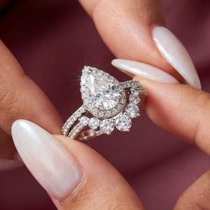 Puede incluir: Un conjunto de anillos de compromiso plateados con una piedra central en forma de pera rodeada por un halo de piedras más pequeñas. La banda está adornada con detalles brillantes adicionales. El anillo se muestra sobre un fondo rosa.