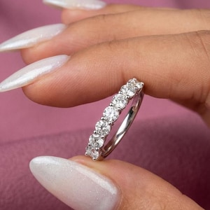 Könnte beinhalten: Ein silberner Ring mit sieben runden, klaren Edelsteinen. Der Ring wird vor einem rosa Hintergrund präsentiert. Die Edelsteine sind in einer Reihe entlang des Bandes angeordnet.