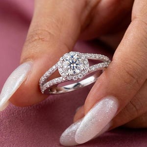 May include: A silver ring with a large, round, clear gemstone surrounded by smaller stones. The ring has a split band, also adorned with small, clear stones. The ring is held against a pink background.