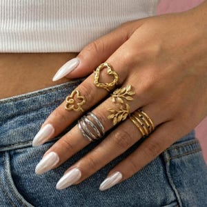 Op de afbeelding: Een hand met verschillende gouden en zilveren ringen. De ringen omvatten een hartvorm, een bloem, een bladontwerp en meerdere banden. De nagels zijn gelakt met een parelmoer witte lak. De achtergrond omvat een witte top en blauwe denim.