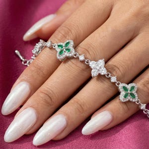 May include: A silver bracelet with clover-shaped designs, set with green and clear gemstones. The bracelet features multiple clover motifs linked by small, sparkling accents. The bracelet is displayed on a pink surface.