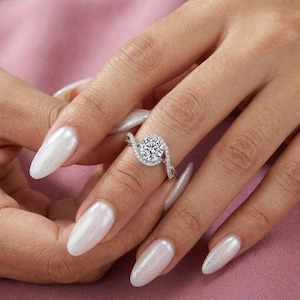 May include: A diamond engagement ring with a round center stone and a halo of smaller diamonds. The ring has a swirling design on the band, and is set on a pink background.