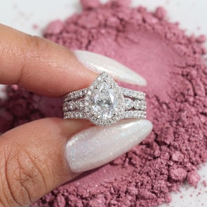 May include: A diamond engagement ring set with a pear-shaped central stone and a halo of smaller diamonds. The ring is displayed against a pink backdrop, highlighting the intricate setting and the sparkle of the gems.