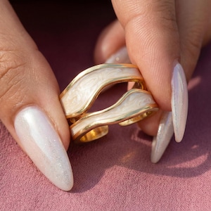 Może przedstawiać: Dwa złote pierścionki o falistym wzorze, z białym emaliowanym wypełnieniem. Pierścionki są trzymane na różowym tle. Pierścionki mają unikalny, nowoczesny design.