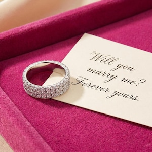 May include: A silver ring with three rows of sparkling stones, presented in a pink velvet box. A card with the words "Will you marry me? Forever yours" is next to the ring, suggesting a proposal.