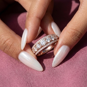 Puede incluir: Anillo de oro rosa con múltiples filas de diamantes redondos brillantes. El anillo presenta una banda de diamantes más pequeños y piedras centrales más grandes. El anillo se sostiene sobre un fondo rosa, mostrando el brillo de la joya.