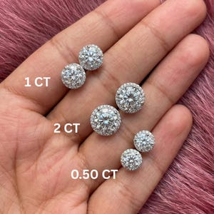 May include: A collection of diamond stud earrings in various sizes, including 0.50 CT, 1 CT, and 2 CT. Each earring features a central diamond surrounded by a halo of smaller stones. The earrings are displayed on a pink surface.
