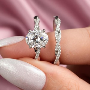 以下が含まれることがあります： 輝く白い宝石が付いた2つのシルバーリング。婚約指輪は大きなオーバルカットの石が特徴で、結婚指輪は小さな石を使ったツイストデザインです。リングはピンク色の背景に保持されています。