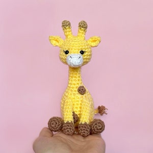 May include: A crocheted yellow giraffe toy with brown spots and a white face. The giraffe is sitting on a hand.