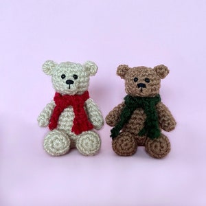 May include: Two crocheted teddy bears wearing scarves. The bear on the left is cream colored and wearing a red scarf. The bear on the right is brown and wearing a green scarf.