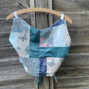 May include: A blue and green patchwork top with a floral and butterfly print. The top has a square neckline and is tied at the back.