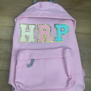 May include: A pink canvas backpack with a large, glittery, multi-coloured patch that reads "HRP".