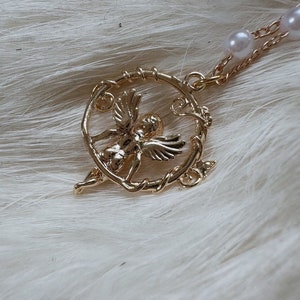 K&ouml;nnte beinhalten: Eine goldene Halskette mit einem Anh&auml;nger, der einen Cherubengel in einem runden Rahmen mit Rankenmuster zeigt. Die Kette besteht aus einer goldenen Kette und hat wei&szlig;e Perlen als Akzente.