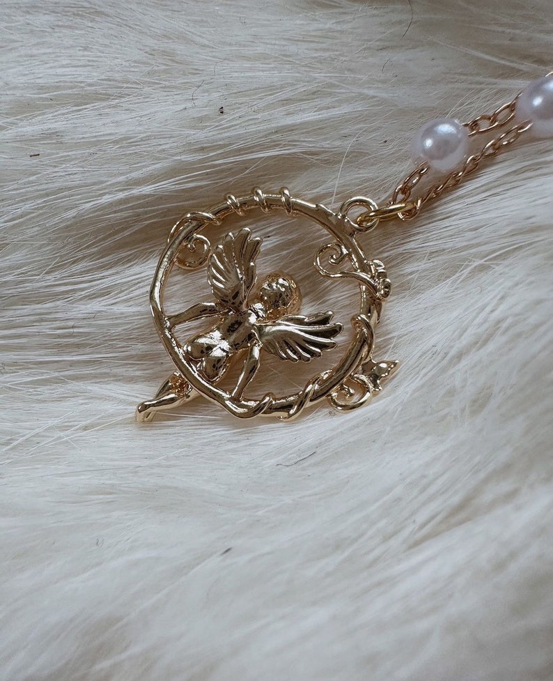K&ouml;nnte beinhalten: Eine goldene Halskette mit einem Anh&auml;nger, der einen Cherubengel in einem runden Rahmen mit Rankenmuster zeigt. Die Kette besteht aus einer goldenen Kette und hat wei&szlig;e Perlen als Akzente.
