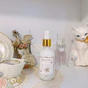 Op de afbeelding: Een witte sprayfles met een gouden kraag en een etiket met de tekst Elegant lace, een hartafbeelding en kleine vlinders. De fles bevat een ondoorzichtige witte vloeistof en is afgesloten met een doorzichtige plastic dop op een witte plank.