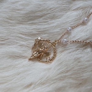 K&ouml;nnte beinhalten: Eine goldene Halskette mit einer zarten Kette und einem Anh&auml;nger mit einem Cherubinenengel in einem runden Rahmen aus goldenen Ranken. Der Anh&auml;nger ist an einer Perlenkette befestigt.