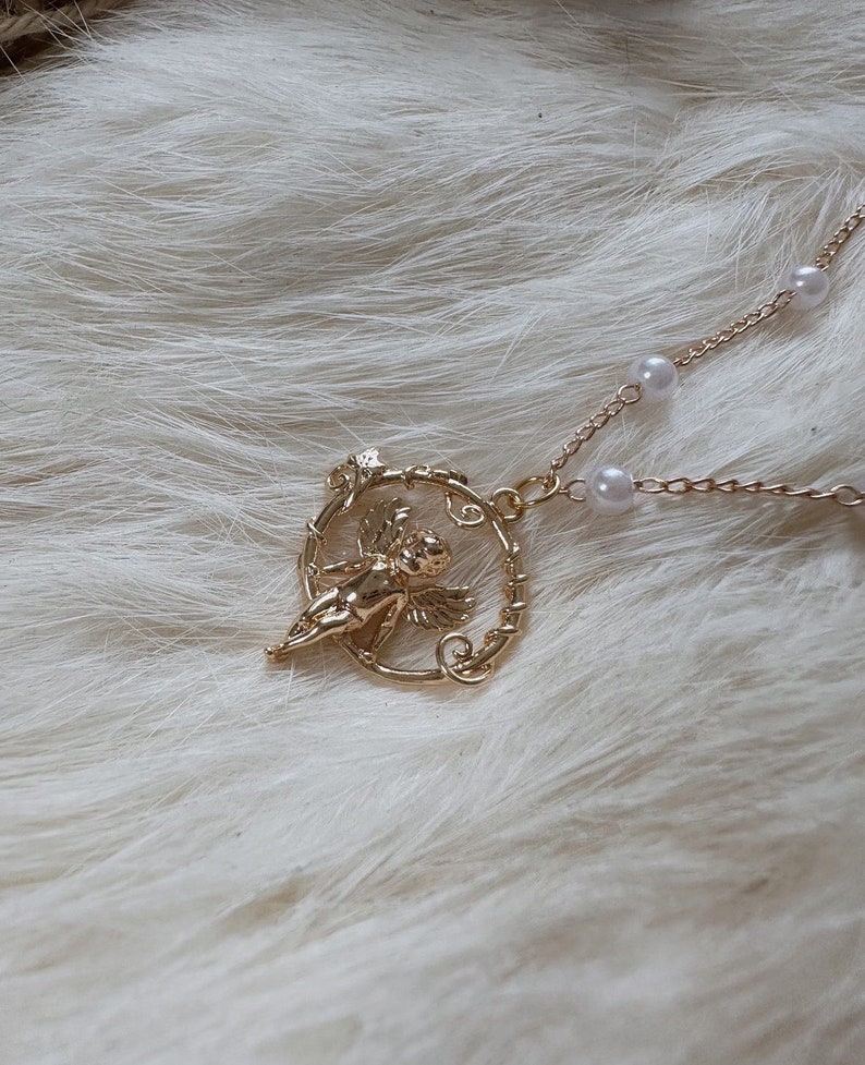 K&ouml;nnte beinhalten: Eine goldene Halskette mit einer zarten Kette und einem Anh&auml;nger mit einem Cherubinenengel in einem runden Rahmen aus goldenen Ranken. Der Anh&auml;nger ist an einer Perlenkette befestigt.