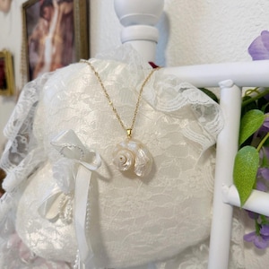 May include: A gold necklace featuring a pearlescent white seashell pendant. The pendant is suspended on a delicate chain. The necklace is displayed on a white lace pillow with a white ribbon and bow. The background includes a white bed frame and a framed picture.