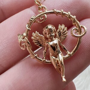 K&ouml;nnte beinhalten: Goldfarbener Engelscharme mit Fl&uuml;geln, der an einem runden, verzierten Rahmen h&auml;ngt. Der Engel ist mit einem cherubischen Gesicht dargestellt und h&auml;lt sich mit seinen H&auml;nden am Rahmen fest.