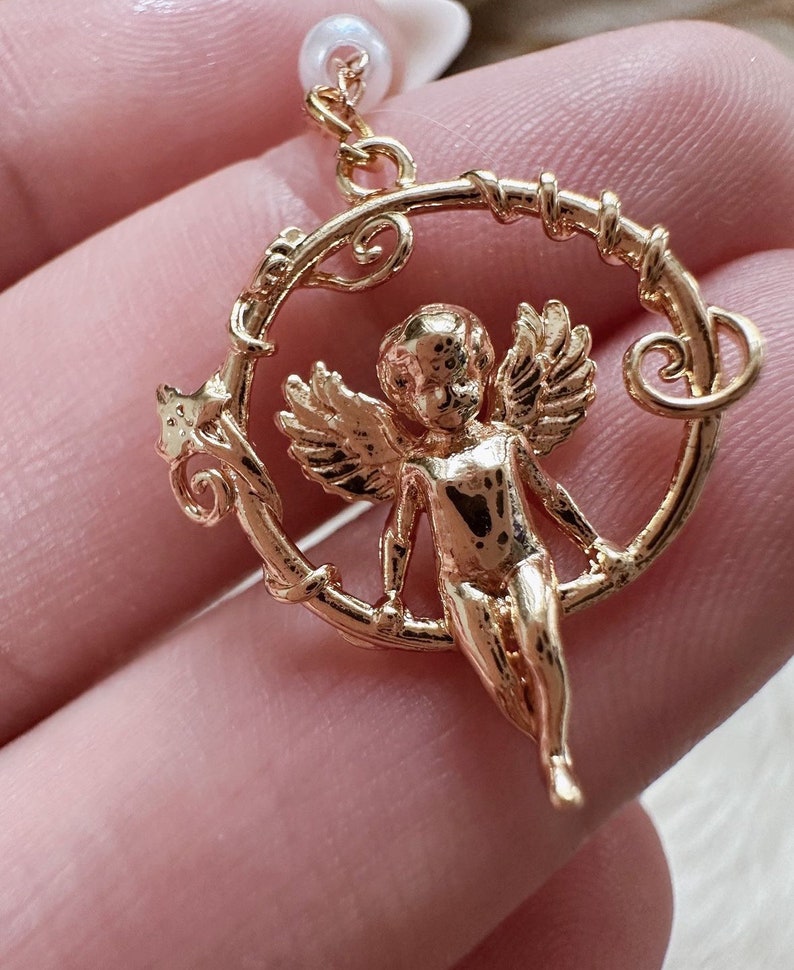 K&ouml;nnte beinhalten: Goldfarbener Engelscharme mit Fl&uuml;geln, der an einem runden, verzierten Rahmen h&auml;ngt. Der Engel ist mit einem cherubischen Gesicht dargestellt und h&auml;lt sich mit seinen H&auml;nden am Rahmen fest.