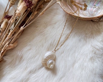 May include: A gold chain necklace with a white seashell pendant. The seashell is a double spiral shape with a pearly finish. The necklace is laying on a white fluffy surface.