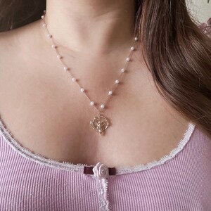 K&ouml;nnte beinhalten: Eine zarte Perlenkette mit einem goldenen Anh&auml;nger, der ein stilisiertes Engelmotiv zeigt. Die Kette wird von einer Person mit langen braunen Haaren und einem hellvioletten Oberteil getragen.