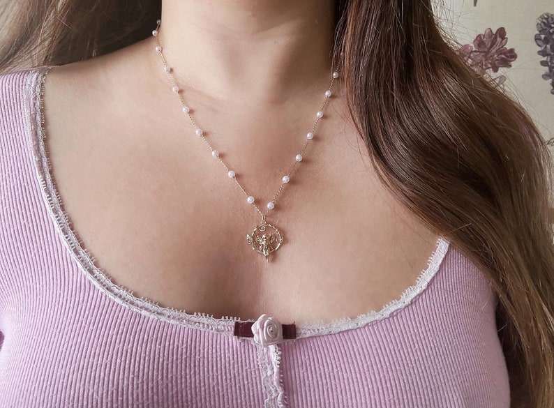 K&ouml;nnte beinhalten: Eine zarte Perlenkette mit einem goldenen Anh&auml;nger, der ein stilisiertes Engelmotiv zeigt. Die Kette wird von einer Person mit langen braunen Haaren und einem hellvioletten Oberteil getragen.