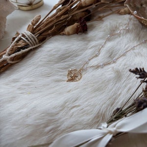 K&ouml;nnte beinhalten: Eine goldene Halskette mit einem zarten, verzierten Anh&auml;nger mit floralem Design. Die Halskette besteht aus kleinen, wei&szlig;en Perlen und wird auf einem wei&szlig;en Kunstfell-Hintergrund pr&auml;sentiert.