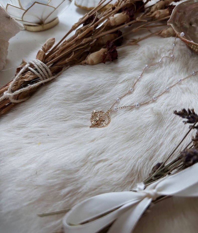 K&ouml;nnte beinhalten: Eine goldene Halskette mit einem zarten, verzierten Anh&auml;nger mit floralem Design. Die Halskette besteht aus kleinen, wei&szlig;en Perlen und wird auf einem wei&szlig;en Kunstfell-Hintergrund pr&auml;sentiert.