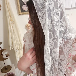Könnte beinhalten: Ein weißer Spitzenschleier mit Rosenmuster. Der Schleier ist über Kopf und Schultern drapiert. Die Person trägt ein weißes Kleid mit rosa Blumenmuster.
