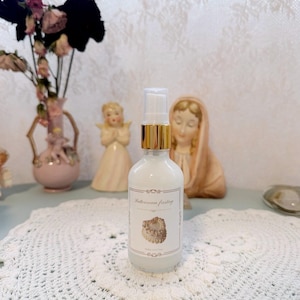 Könnte beinhalten: Eine durchsichtige Glassprühflasche gefüllt mit einer weißen Flüssigkeit, mit einem goldfarbenen Kragen und einem weißen Pumpkopf. Das Etikett zeigt den Text Buttercream frosting und eine Abbildung eines kleinen, runden Kuchens mit weißer Glasur.