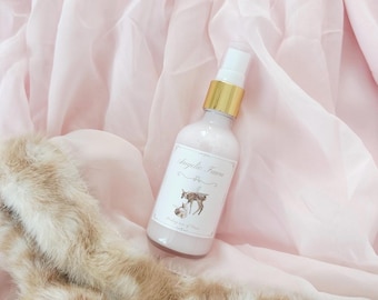 May include: A clear plastic spray bottle filled with a pale pink liquid, featuring a gold-colored pump top. The white label displays the text Angelic Fawn above an illustration of a small deer and a rabbit, with additional small text below.