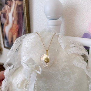 Op de afbeelding: Een ketting met een hartvormige hanger en een gouden ketting. De hanger is wit met een gouden zonnegelaat ontwerp. De ketting wordt gepresenteerd op een wit, met kant bedekt kussen. De achtergrond omvat een ingelijst kunstwerk en een wit decoratief element.
