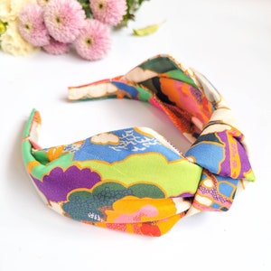 May include: A colorful fabric headband with a knotted design. The headband features a vibrant pattern of green, blue, purple, orange, and cream-colored clouds and landscapes. The accessory is designed for hair styling and fashion.