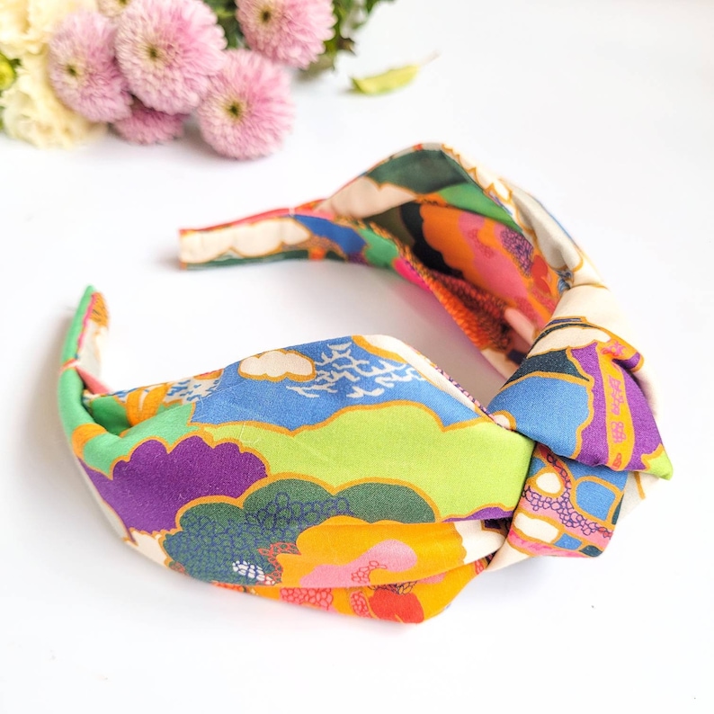May include: A colorful fabric headband with a knotted design. The headband features a vibrant pattern of green, blue, purple, orange, and cream-colored clouds and landscapes. The accessory is designed for hair styling and fashion.