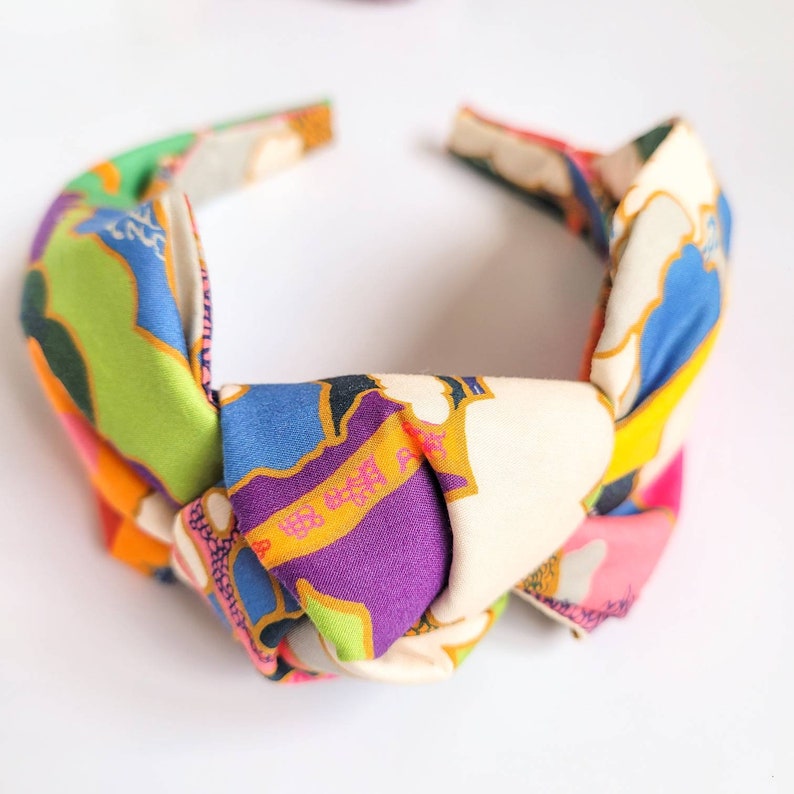 May include: A colorful knotted headband with a vibrant floral pattern. The headband features a mix of colors, including green, blue, purple, pink, and orange, creating a bold and eye-catching accessory. The fabric has a silky sheen.