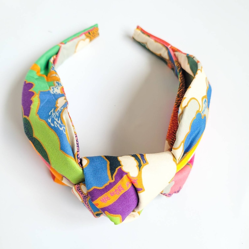 May include: A colorful fabric headband with a knotted design. The headband features a vibrant mix of colors, including green, blue, purple, orange, and pink, with floral and abstract patterns. The accessory is designed to be worn on the head.