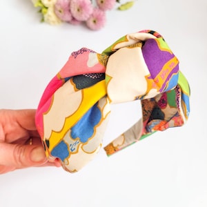 May include: A colorful knotted headband with a vibrant floral pattern. The headband features a mix of pink, yellow, purple, and green hues. The fabric has a silky sheen, and the design is a mix of abstract shapes and floral motifs.
