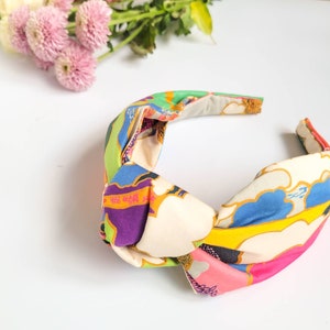 May include: A colorful knotted headband with a vibrant pattern. The headband features a cream-colored base with a design of blue, green, purple, and pink shapes. The fabric is knotted at the top, creating a stylish accessory.