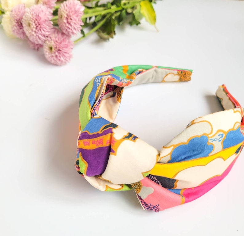 May include: A colorful knotted headband with a vibrant pattern. The headband features a cream-colored base with a design of blue, green, purple, and pink shapes. The fabric is knotted at the top, creating a stylish accessory.