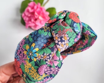 May include: A floral print headband with a knotted top. The fabric features a navy blue background with a colorful pattern of flowers in shades of pink, yellow, green, and blue. The headband is held by a hand.