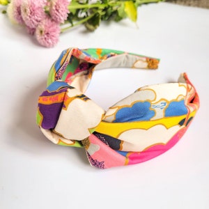 May include: A colorful knotted headband with a vibrant floral and cloud pattern. The headband features a mix of pink, blue, green, yellow, and purple hues. The fabric is knotted at the top, creating a stylish accessory.