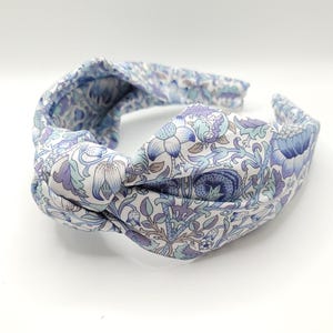 May include: A fabric headband with a knotted design. The headband features a floral pattern in shades of blue, purple, and green on a white background. The headband is a hair accessory.