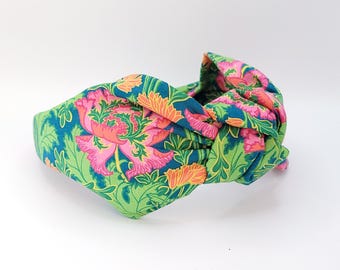 May include: A fabric headband with a knotted top. The headband features a floral pattern with pink, orange, and green colors. The base color is green with a floral design in pink and orange.