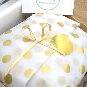 May include: A gift wrapped in white paper with gold polka dots, tied with a gold ribbon. A gold sticker and a card with the text "Dolores Apple Handmade Crafts" are visible.