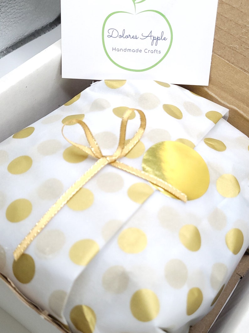 May include: A gift wrapped in white paper with gold polka dots, tied with a gold ribbon. A gold sticker and a card with the text "Dolores Apple Handmade Crafts" are visible.