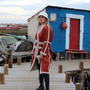 May include: A woman wearing a red and white shearling coat with a white bucket hat, black pants, and black boots. She is standing on a wooden dock in front of a blue and red building.