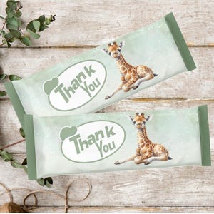 May include: Two light green candy bar wrappers with a watercolour effect, featuring a baby giraffe illustration and the words "Thank You" in a white oval. The wrappers have a green border and are on a wooden surface.
