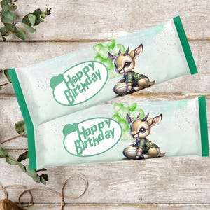 Puede incluir: Dos envoltorios de caramelos verdes y blancos con un ciervo de dibujos animados con un suéter verde. Los envoltorios tienen el texto "Happy Birthday" en un óvalo verde. Los envoltorios están sobre una superficie de madera.