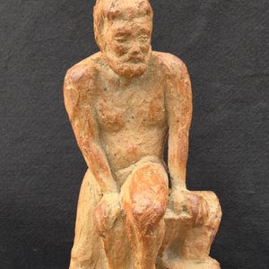 May include: A terracotta-colored clay sculpture of a seated figure with a beard, depicted in a contemplative posture. The figure has a textured, earthy surface with visible hand-molded details, showing a person resting with hands on knees.